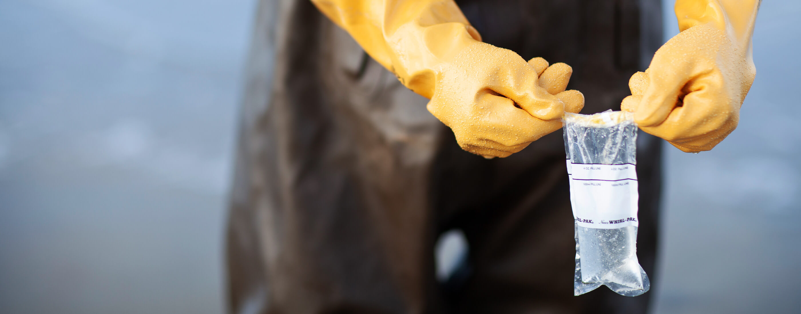 Hands in yellow safety gloves hold up filled water sample bag on the beach.